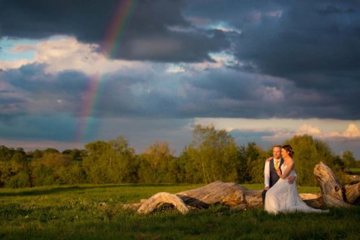 bride and groom sitting together on the tree trunk on the field with a rainbow behind them at winkworth farm