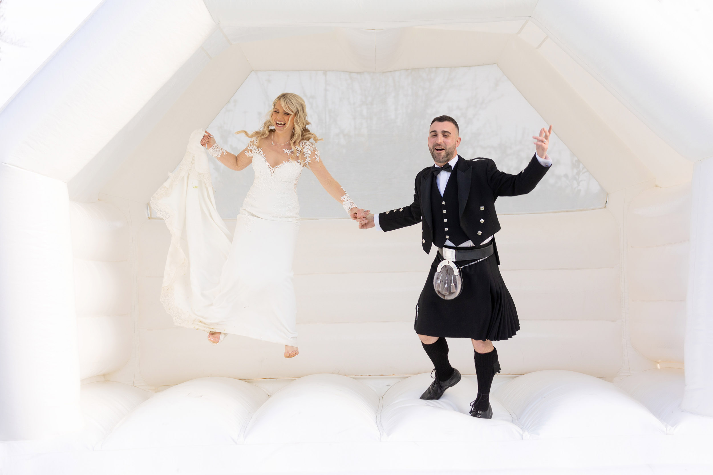 bride and groom in kilt jumping on bouncy castle holding hands