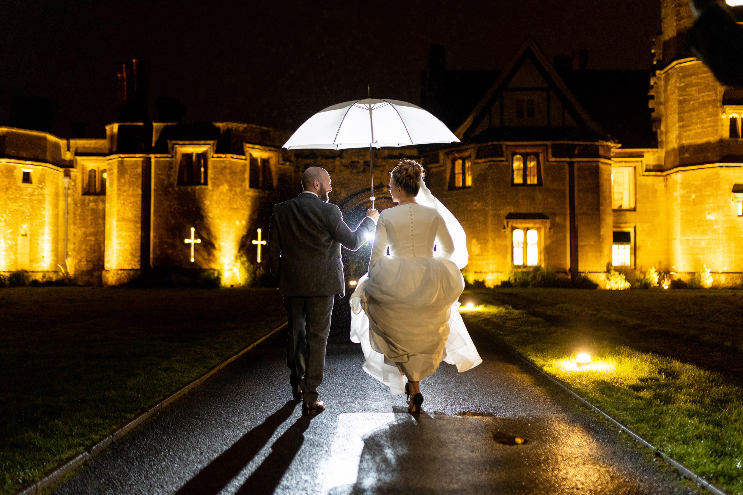 bride and groom walking towards thornbury castle at night holding white umbrella