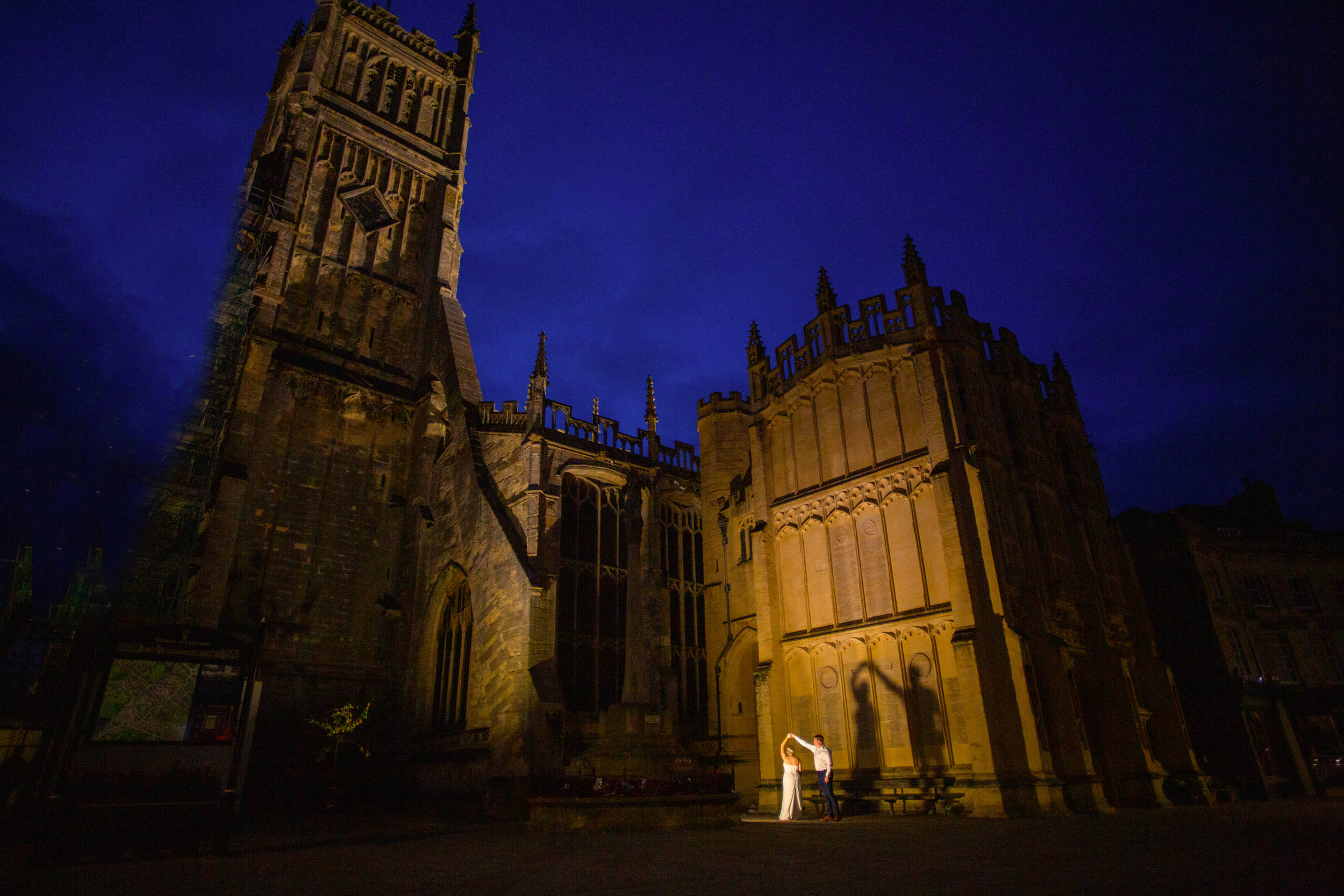 BRIDE AND GROOM DANCING AT NIGHT IN FRONT OF CIRENCESTER CATHEDRAL CASTING THEIR SHADOW ON THE BUILDING