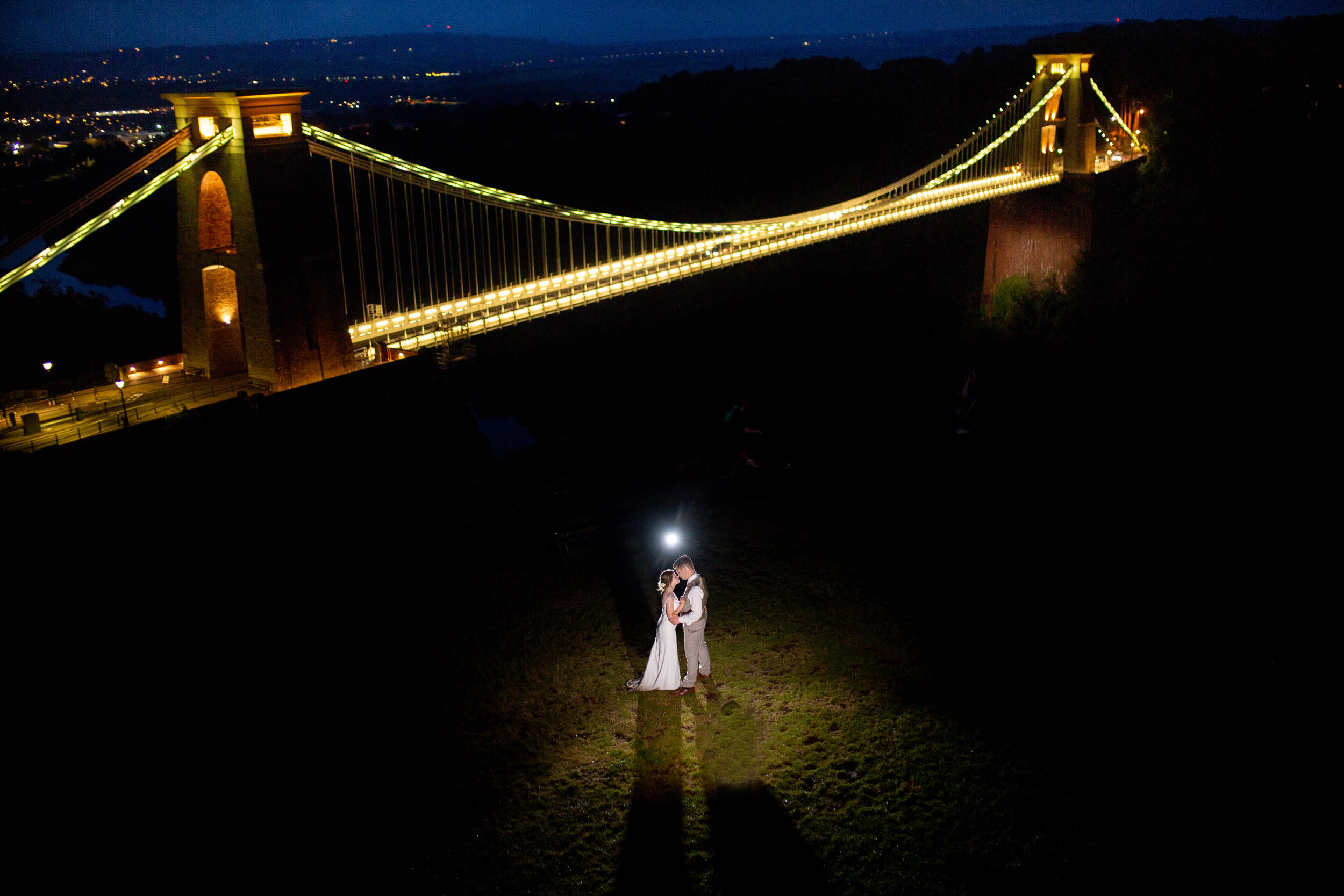 BRIDE AND GROOM KISSING EACH OTHER AT NIGHT WITH LIT CLIFTON SUSPENSION BRIDGE IN THE BACKGROUND. CLIFTON OBSERVATORY WEDDING PHOTOGRAPHY