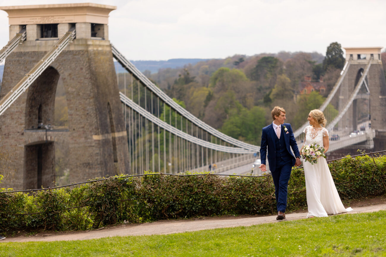 BRIDE AND GROOM WALKING HOLDING HANDS WITH THE CLIFTON SUSPENSION BRIDGE IN THE BACKGROUND